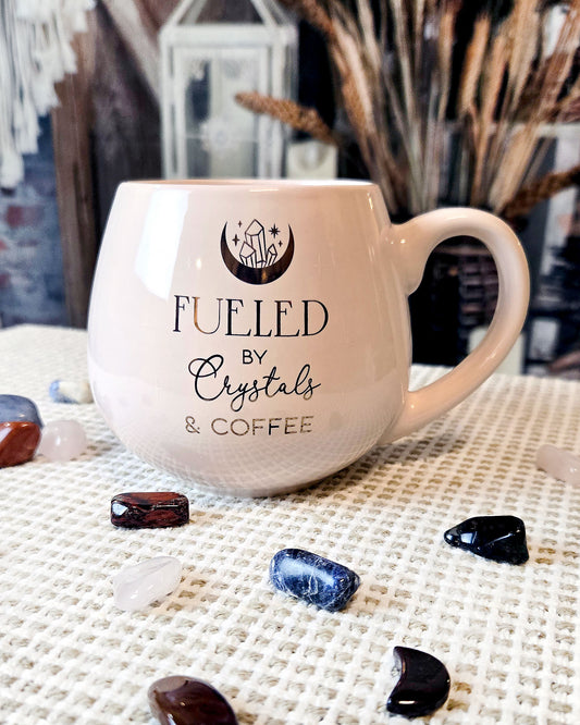 light pink coffee mug that says fueled by crystals and coffee with moon and crystal design in gold front